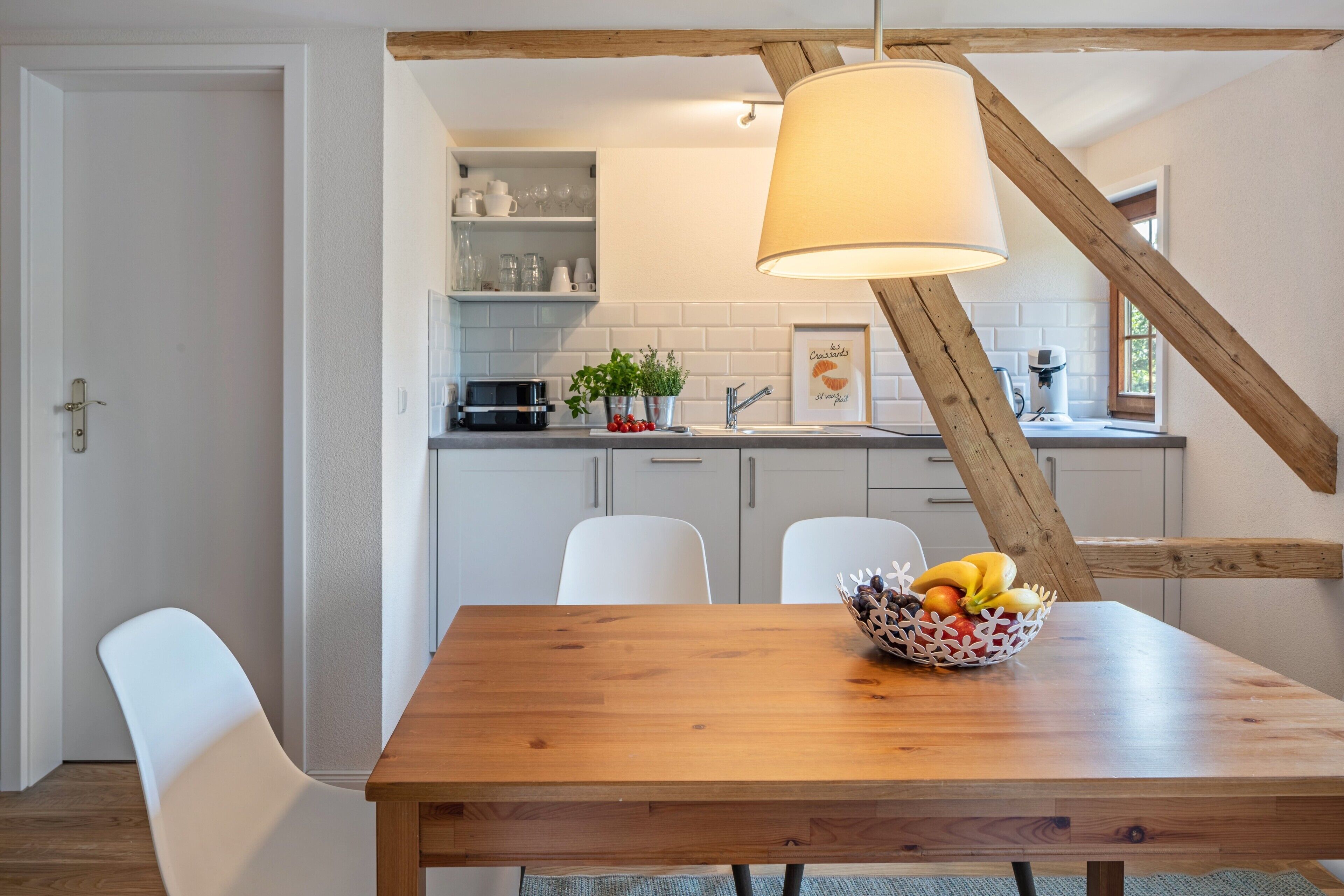 A dining area with a wooden table, white chairs, a fruit bowl, and a kitchen in the background with white cabinets and a hanging light fixture.