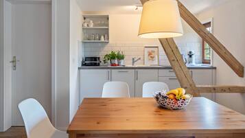 A dining area with a wooden table, white chairs, a fruit bowl, and a kitchen in the background with white cabinets and a hanging light fixture.