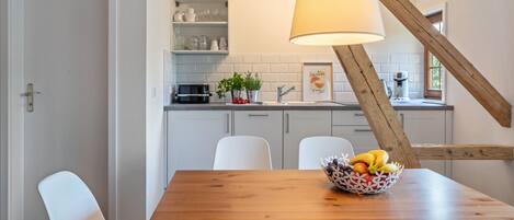 A dining area with a wooden table, white chairs, a fruit bowl, and a kitchen in the background with white cabinets and a hanging light fixture.
