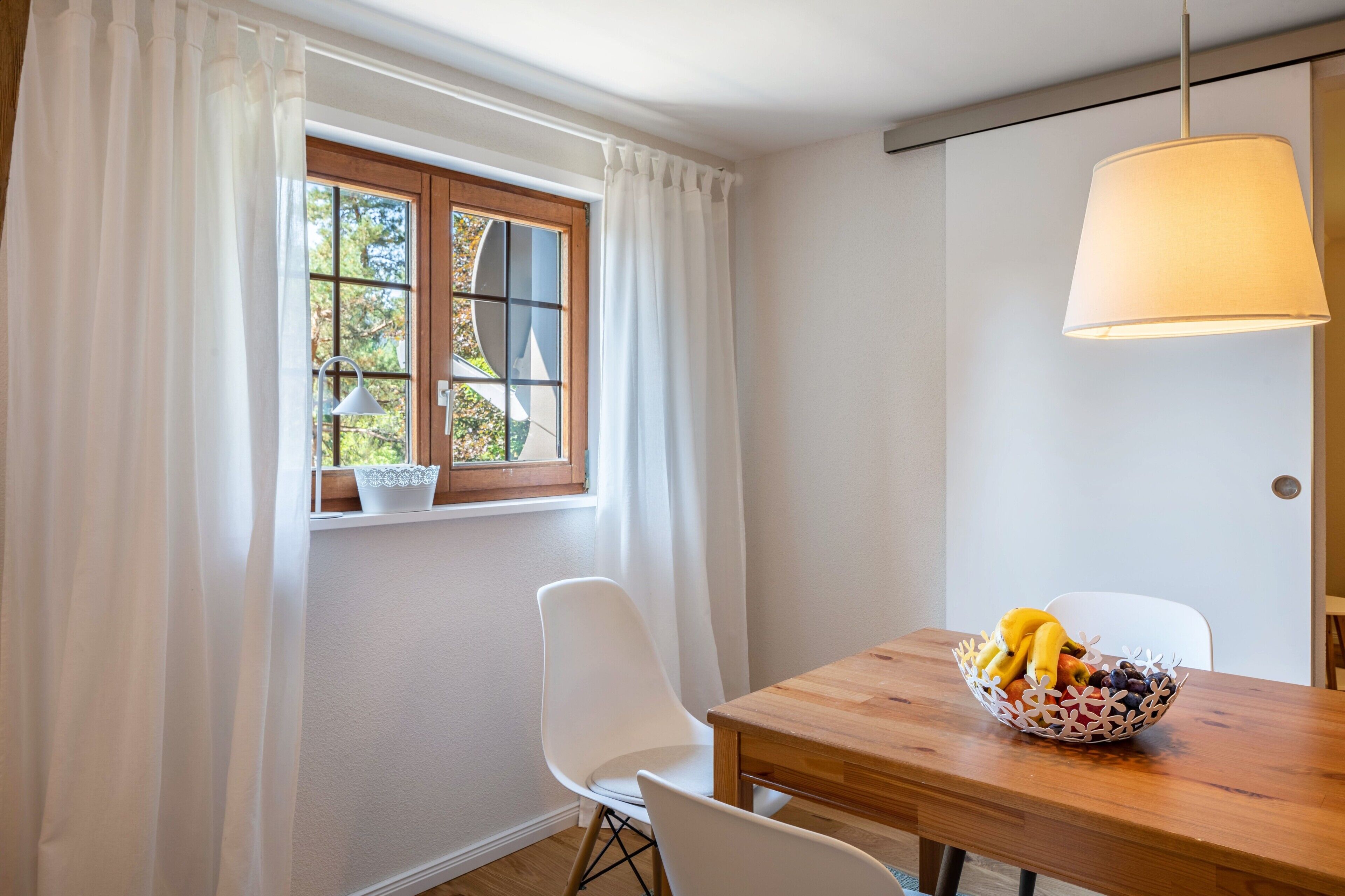 Ein Essbereich mit einem Holztisch, weißen Stühlen, einer Obstschale und einem Fenster mit Vorhängen.