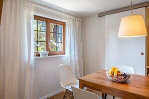 Ein Essbereich mit einem Holztisch, weißen Stühlen, einer Obstschale und einem Fenster mit Vorhängen.