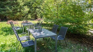 Et udendørs opholdsområde med metalborde og stole, omgivet af frodig vegetation og træer.