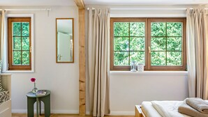 Een slaapkamer met een houten bed, een klein nachtkastje met een vaas en boek, een raam met gordijnen en een spiegel aan de muur.