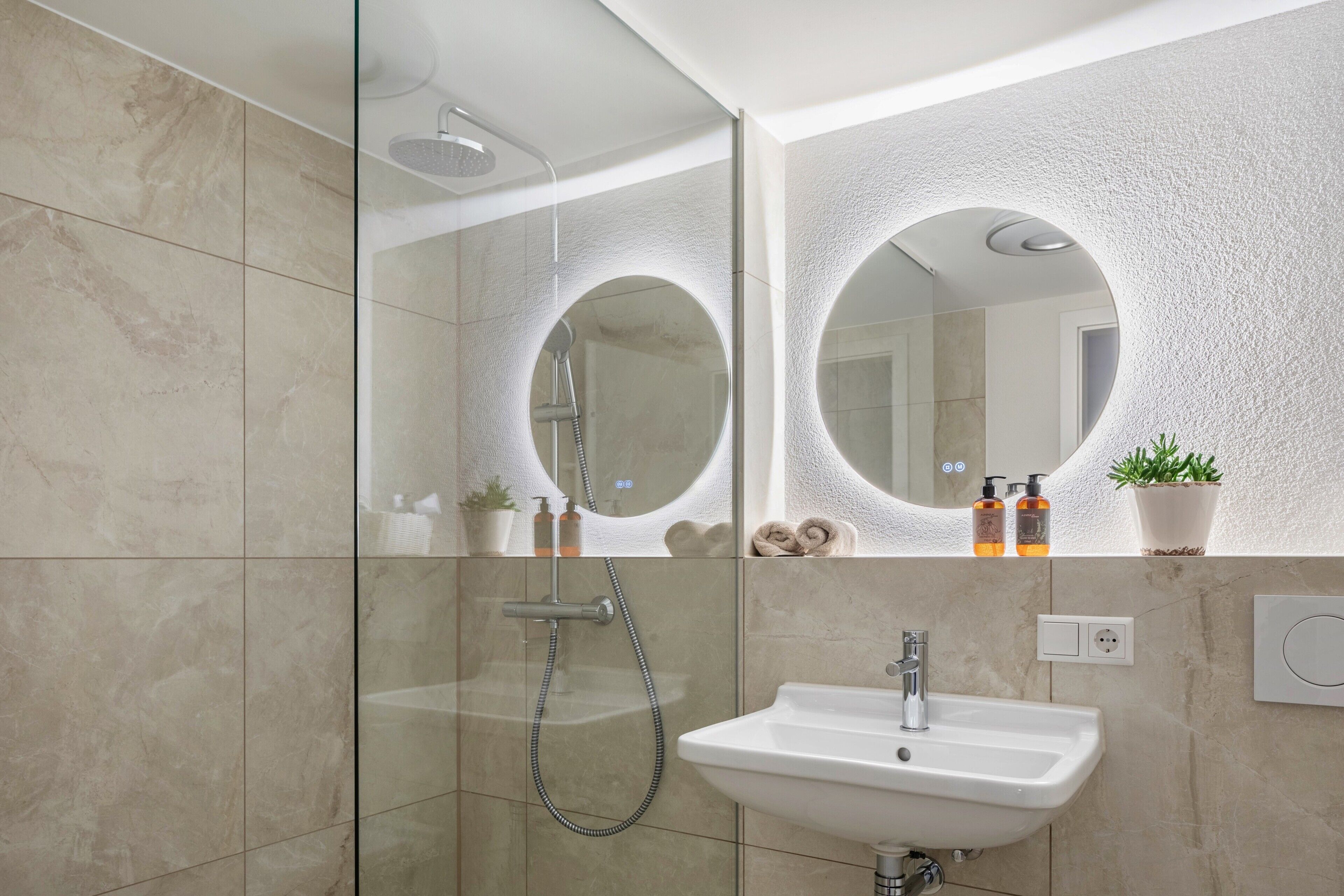 Ein modernes Badezimmer mit einer gläsernen Duschkabine, einem weißen Waschbecken und einem großen Spiegel an einer strukturierten Wand.