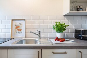 Une cuisine avec un évier en acier inoxydable, un tableau représentant des croissants, une plante en pot et une planche à découper avec des tomates.