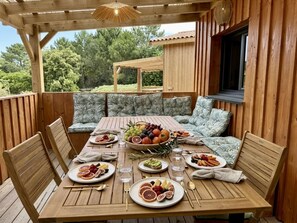 An outdoor wooden deck with a table set for a meal, surrounded by cushioned seating. - Chalet 76 ⭐️ Terrace ⭐️ Sleeps 6 ⭐️ Beach ⭐️ A/C (Vendays-Montalivet)
