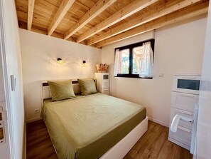 A bedroom with a wooden ceiling, a bed with green bedding, two wall-mounted lights, a window with a curtain, and a small white cabinet. - Chalet 76 ⭐️ Terrace ⭐️ Sleeps 6 ⭐️ Beach ⭐️ A/C (Vendays-Montalivet)