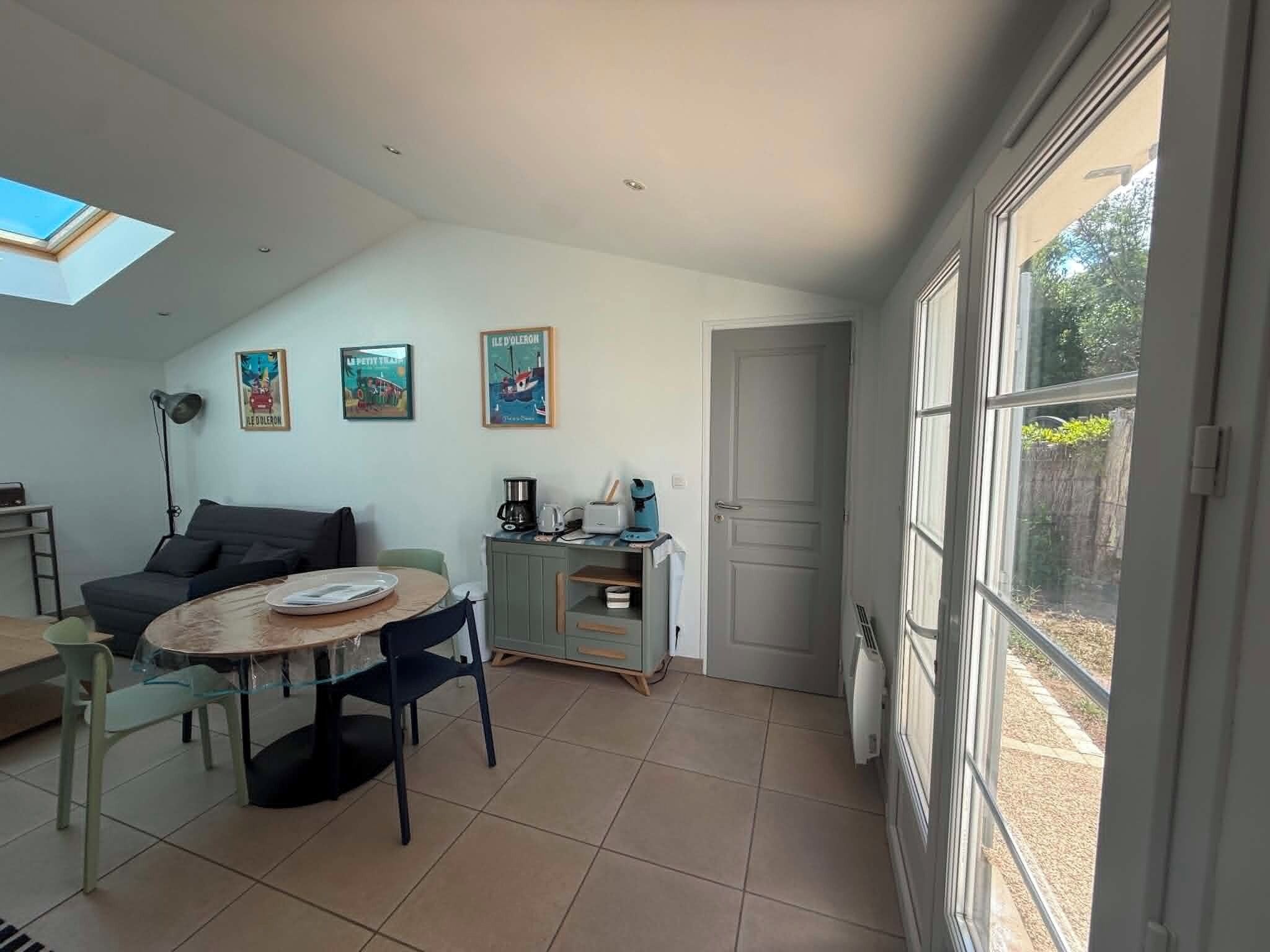 Ein stilvolles Wohnzimmer mit Essbereich, einer Couch, einem Couchtisch und einem Schrank. An der Wand hängen drei gerahmte Bilder und ein großes Fenster bietet Blick ins Freie.