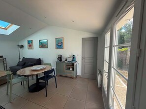 Ein stilvolles Wohnzimmer mit Essbereich, einer Couch, einem Couchtisch und einem Schrank. An der Wand hängen drei gerahmte Bilder und ein großes Fenster bietet Blick ins Freie.