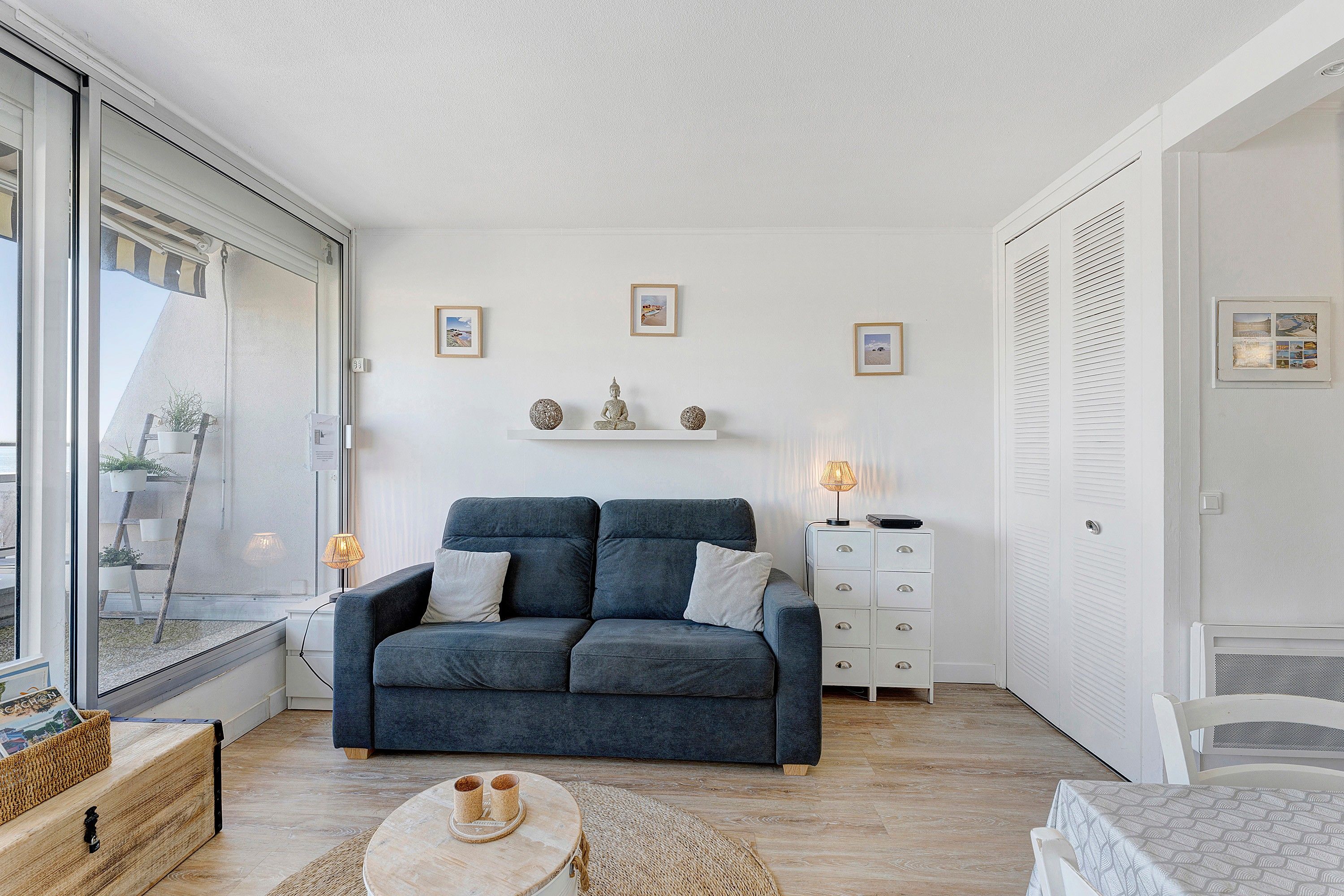 A modern living room with a grey sofa, a small round coffee table, and a white cabinet with drawers. There are framed pictures on the wall and a window with a view of the outside.