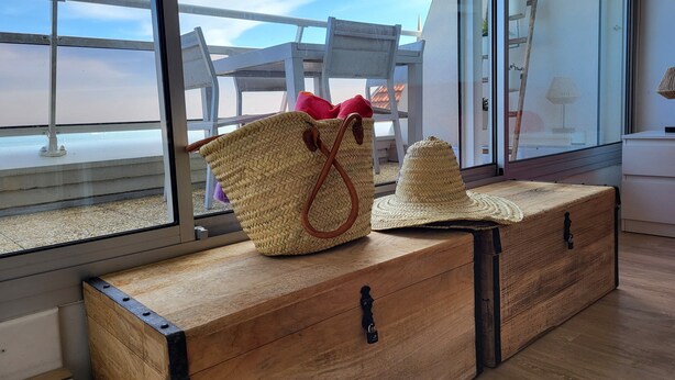 A wooden chest with a woven bag and a hat on top, placed near a window with a view of outdoor furniture. - Apartment 'Carpe Diem' with Bassin View, Balcony and Wi-Fi (Arcachon)