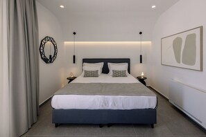 A modern bedroom with a large bed, bedside tables, a wall-mounted light fixture, and a framed abstract painting. - Trinity Secret Villas (fira)