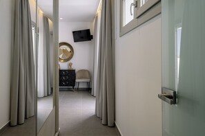 A hallway with a door, a TV mounted on the wall, a round mirror, a chair, and a dresser. - Trinity Secret Villas (fira)