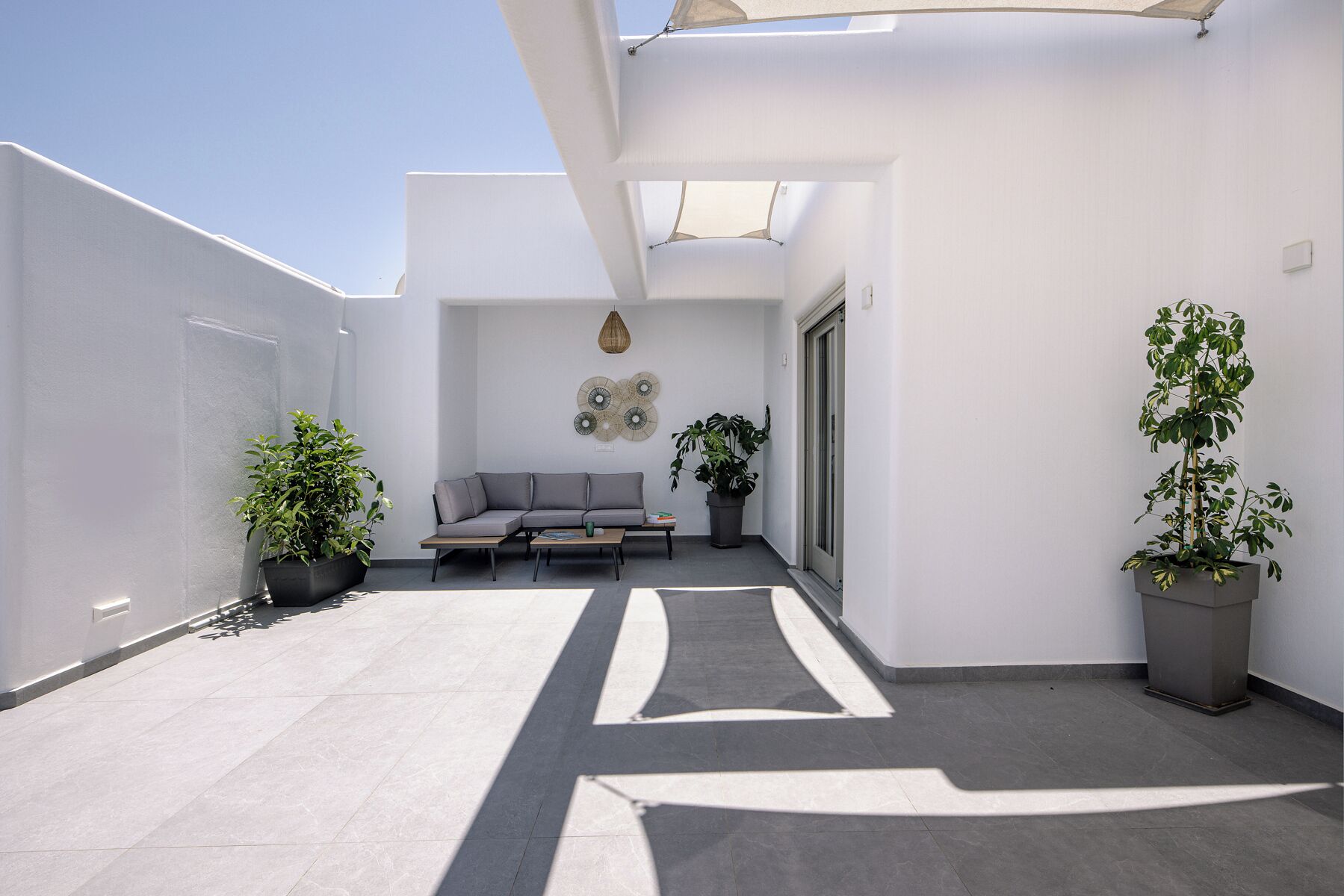 Un patio moderno al aire libre con un sofá gris, dos plantas en macetas y un adorno de pared decorativo.
