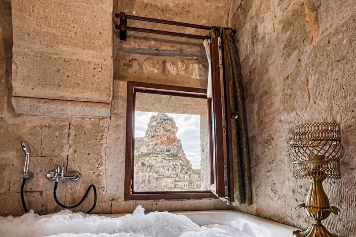 Ein rustikales Badezimmer mit Steinwand, einem Fenster mit Bergblick, einer mit Blasen gefüllten Badewanne und einer dekorativen Lampe.