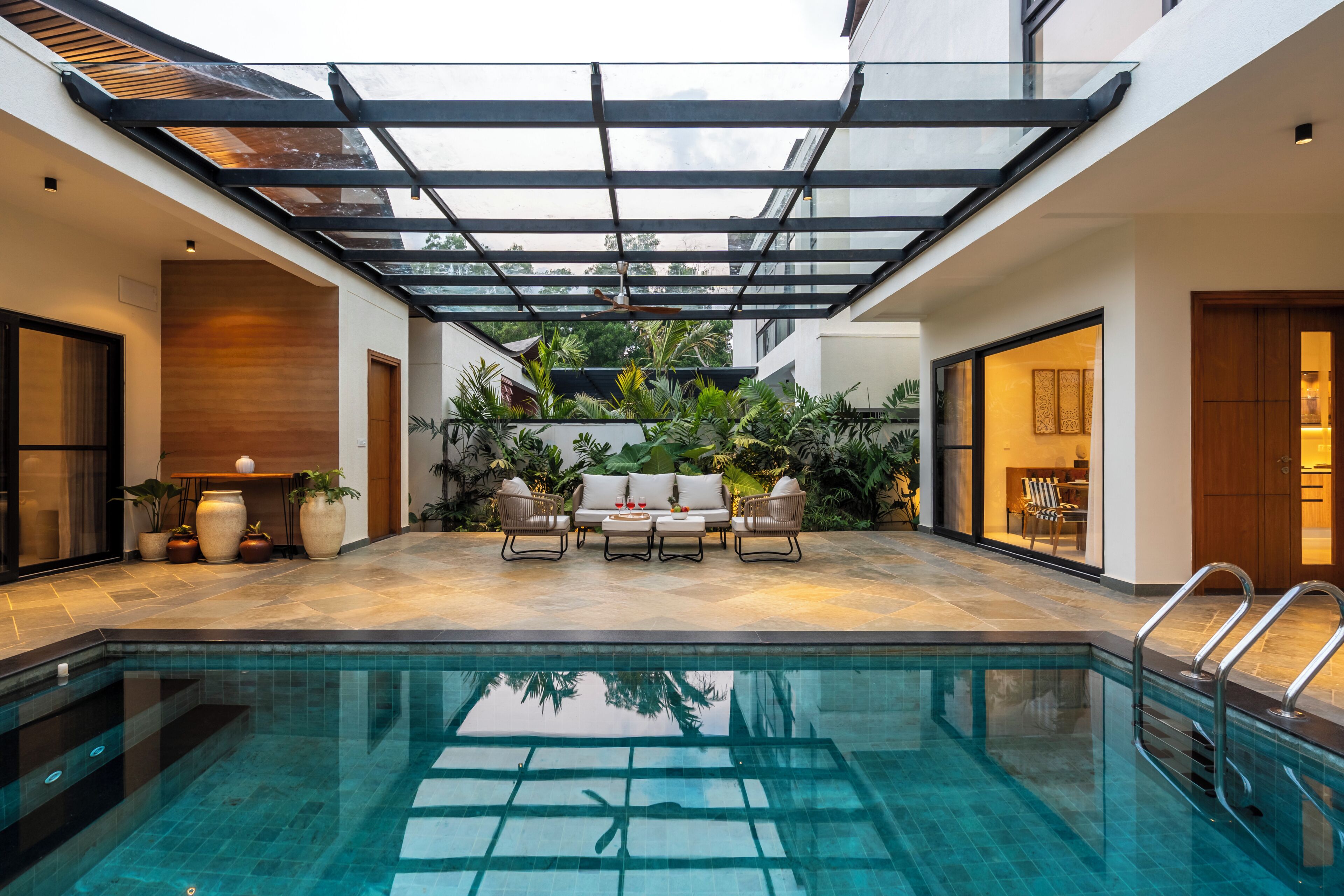 A modern indoor pool area with a glass ceiling, a seating area with a sofa and chairs, and a pool with clear blue water.