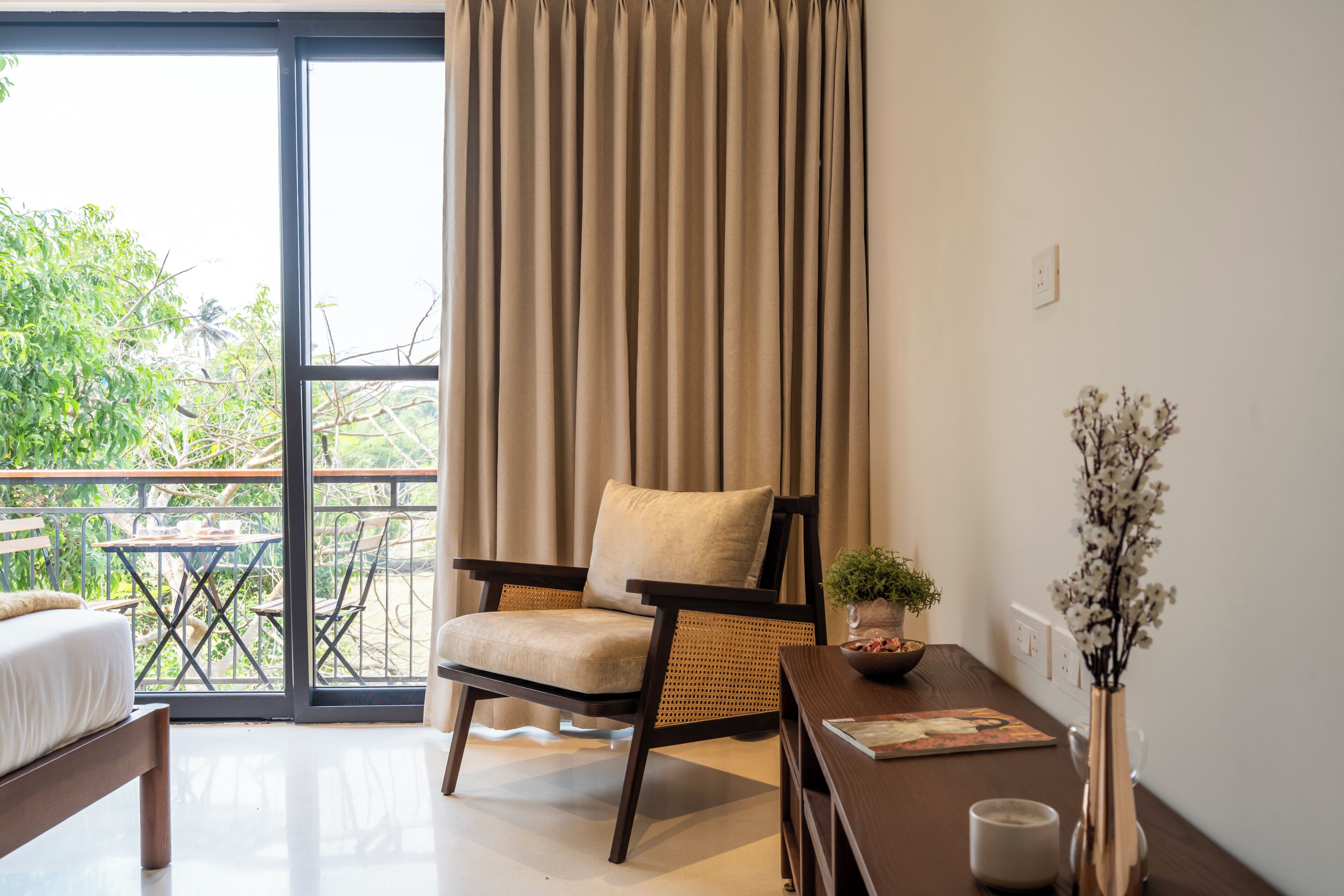 A room with a bed, a chair, a table, a vase with flowers, and a view through a large window.