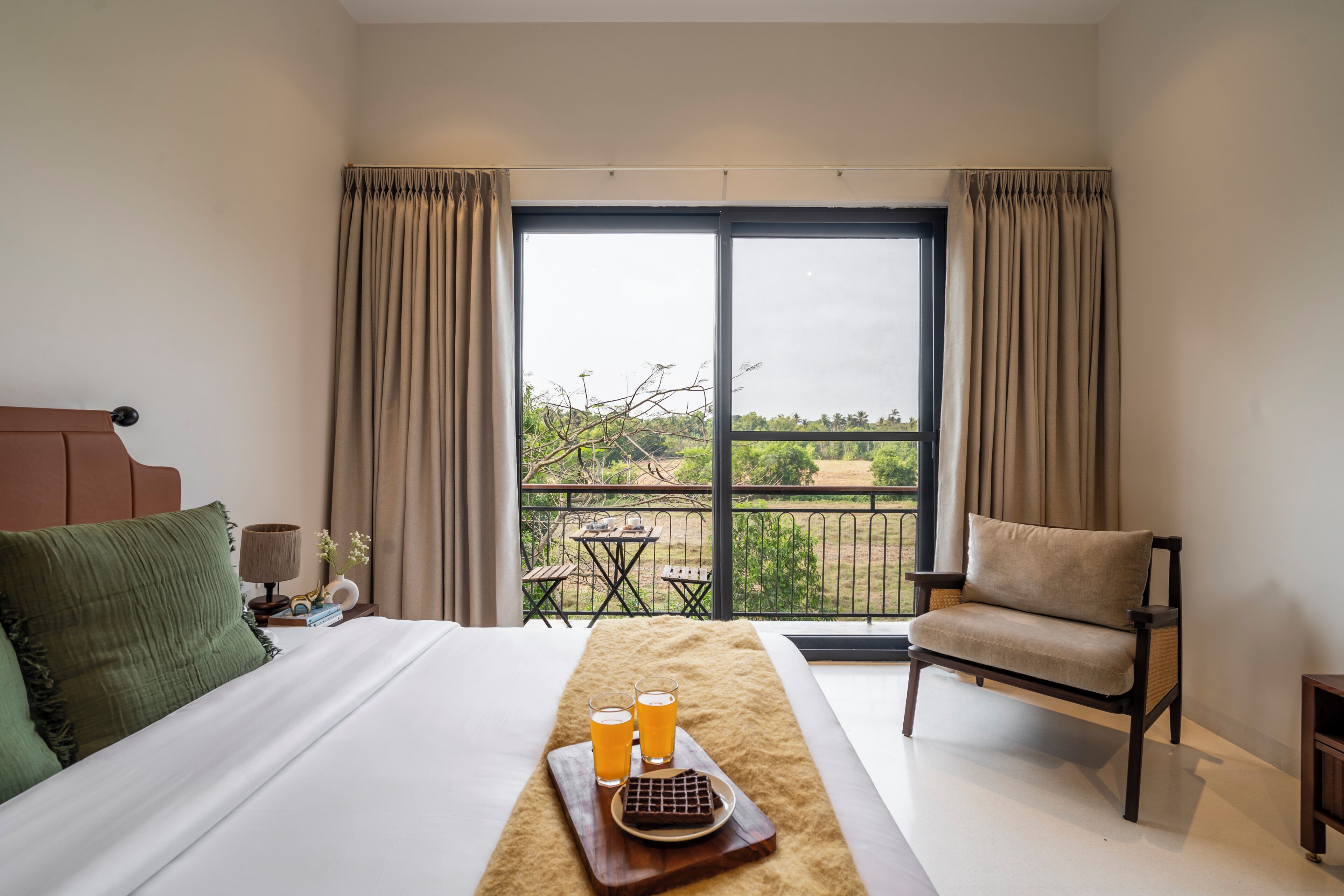 A hotel room with a bed, a chair, a balcony with a table, and a view of greenery.