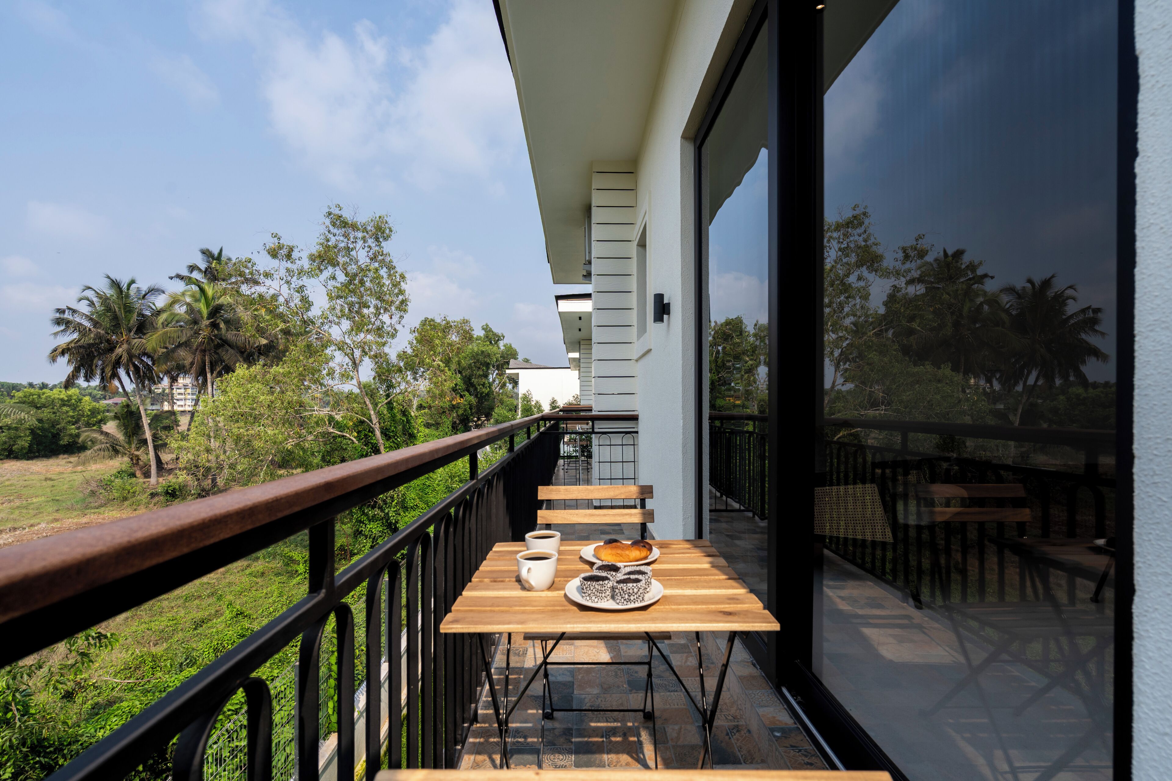 Un balcón con una mesa lista para el desayuno, barandilla y vistas a zonas verdes y edificios.