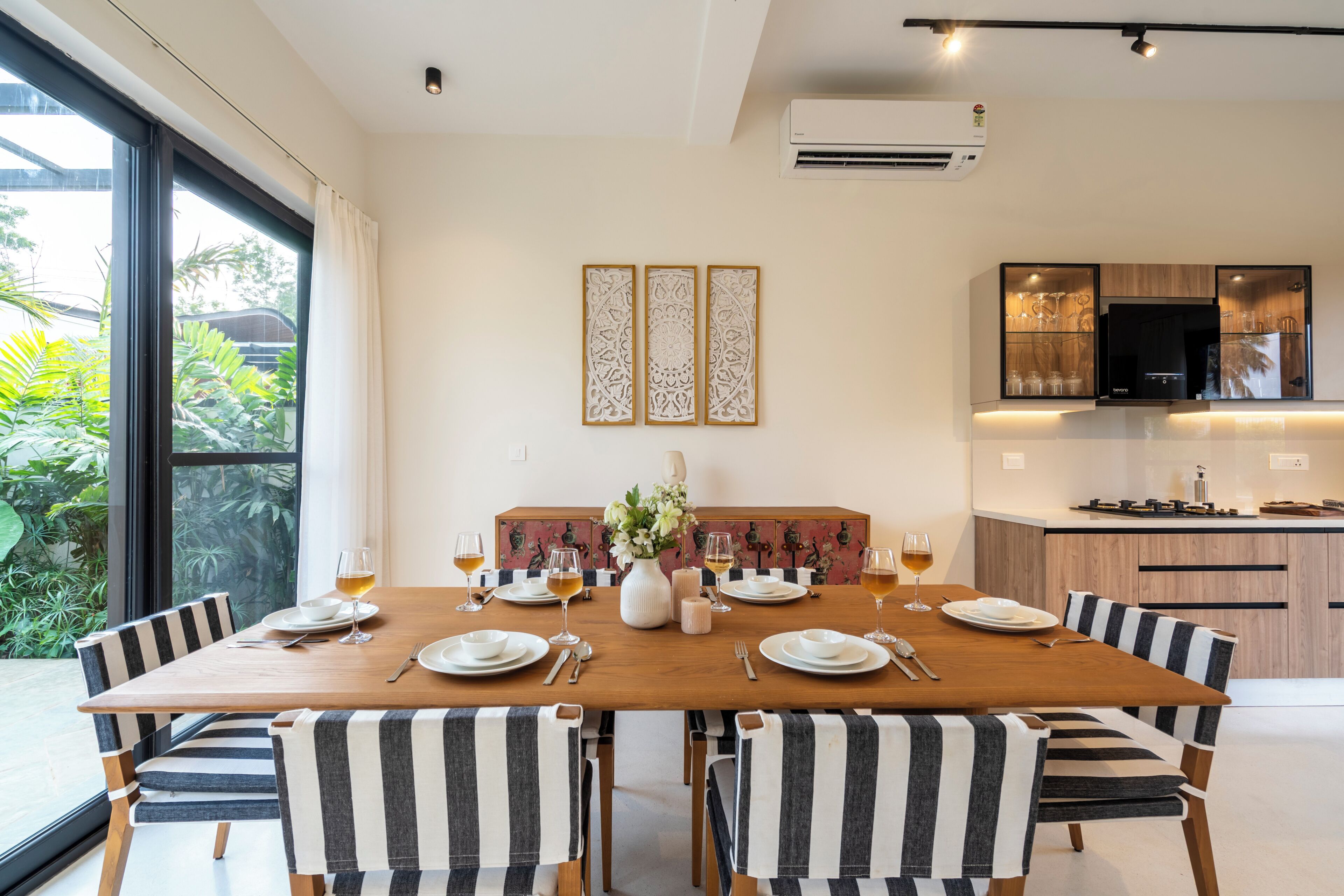 A dining area with a wooden table set for four, surrounded by striped chairs, with a view of greenery outside.