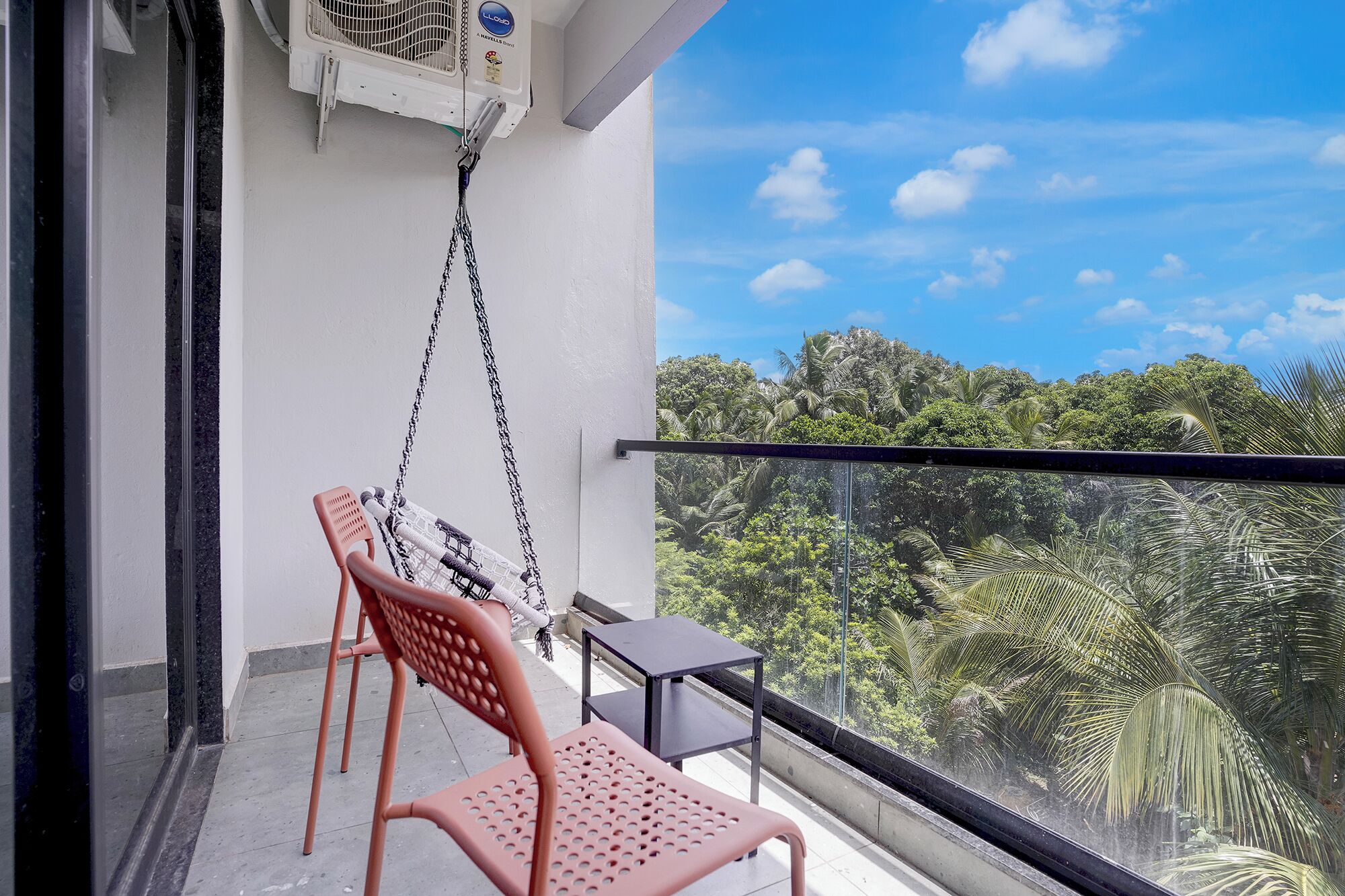 A balcony with a hanging chair, a small table, and a view of dense greenery.