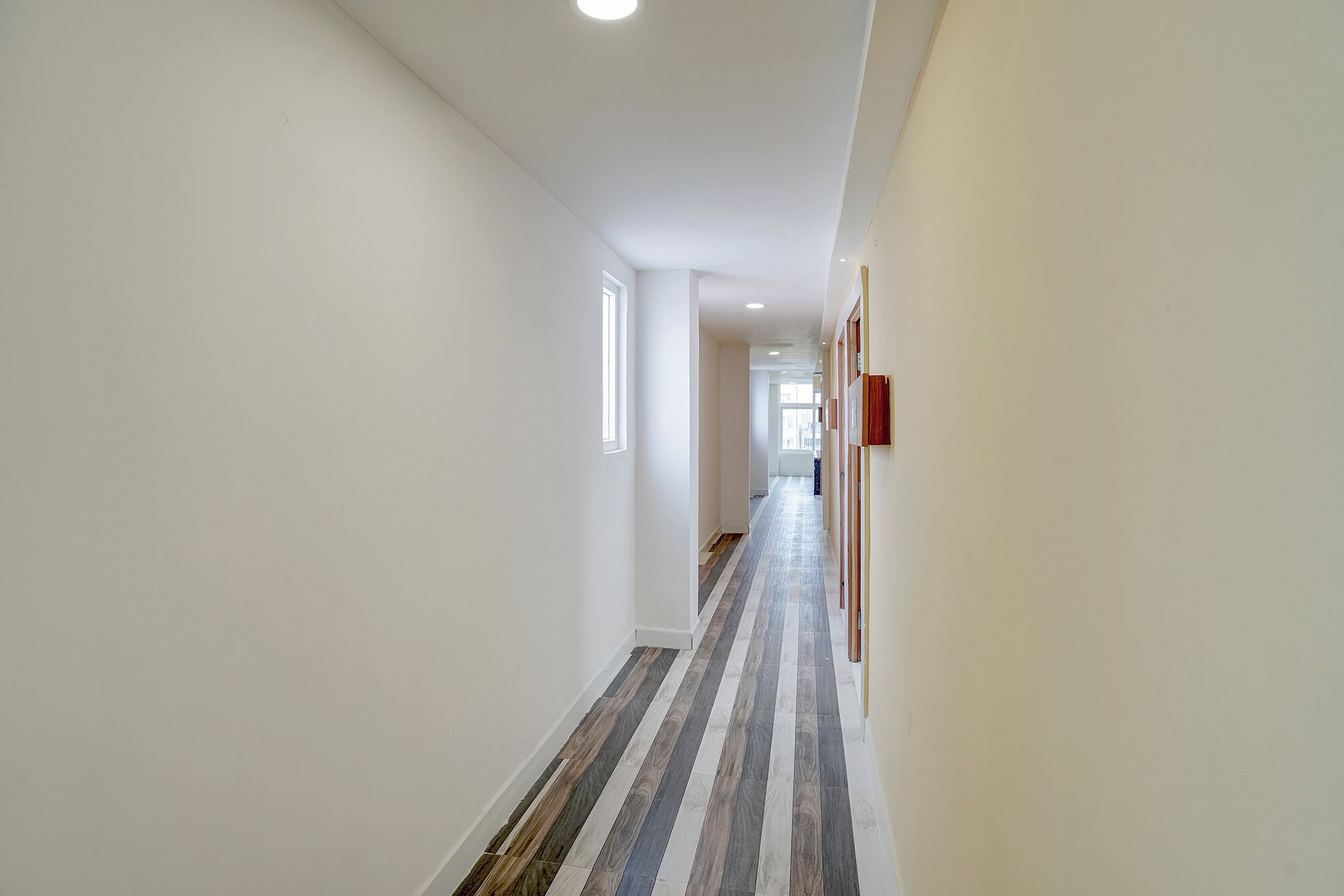 A long, narrow hallway with a striped floor, white walls, and a ceiling with recessed lighting.