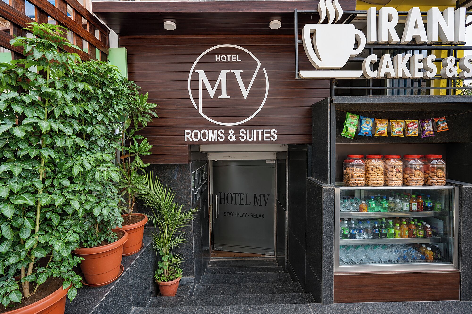 Entrada de un hotel con un cartel que dice 'Hotel MV Rooms & Suites' y una tienda de 'Cakes & Sweets'.