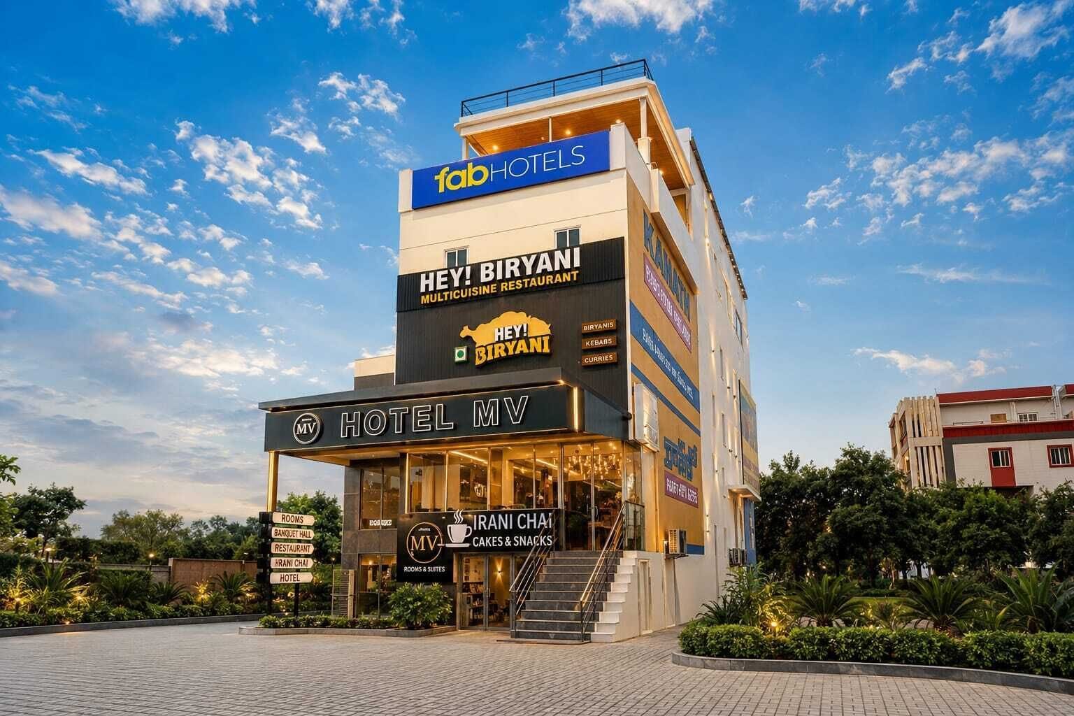 Et hotel med flere etager, med et skilt for 'Hotel MV' og en restaurant, der hedder 'Hey! Biryani Multicuisine Restaurant', på øverste etage.