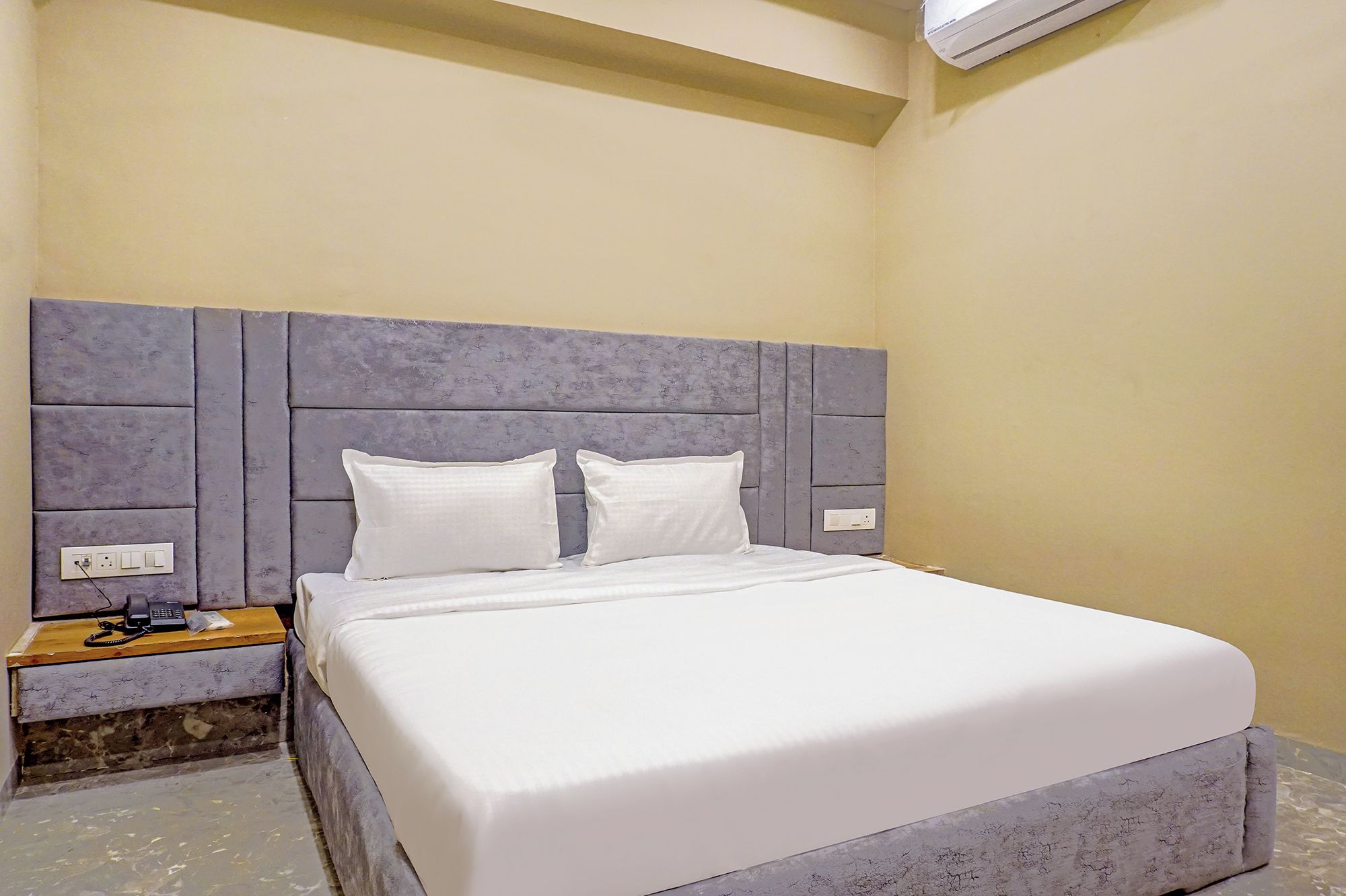A hotel room with a large bed, a headboard with a textured design, a bedside table with a phone, and an air conditioning unit mounted on the wall.