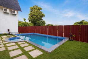 A rectangular swimming pool with a tiled edge, surrounded by a red fence, and a grassy area with stepping stones leading to the pool. - FabHotel Hill View Cottages (Arpora)