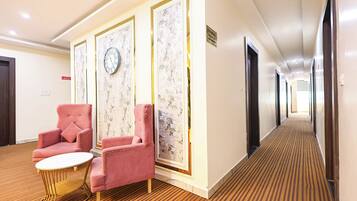 Un pasillo de hotel con piso alfombrado, un reloj en la pared y dos sillas rosas con una mesita.