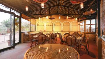 Un interior de restaurante con mesas redondas, sillas de mimbre y lámparas decorativas en el techo.