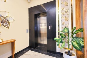 Un ascensor con pantalla digital, una cámara de seguridad en la pared y una planta en maceta de hojas variegadas.