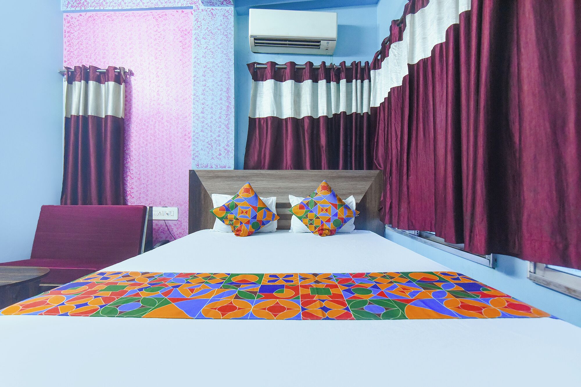 A hotel room with a bed, two pillows, a headboard, a window with curtains, an air conditioning unit, and a wall with a textured pattern.