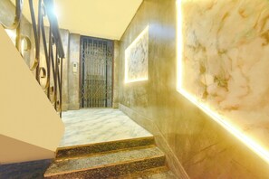 Un escalier en marbre, surmonté d’une grille métallique, mène à une pièce dont un mur est également en marbre et où est accroché un tableau encadré.
