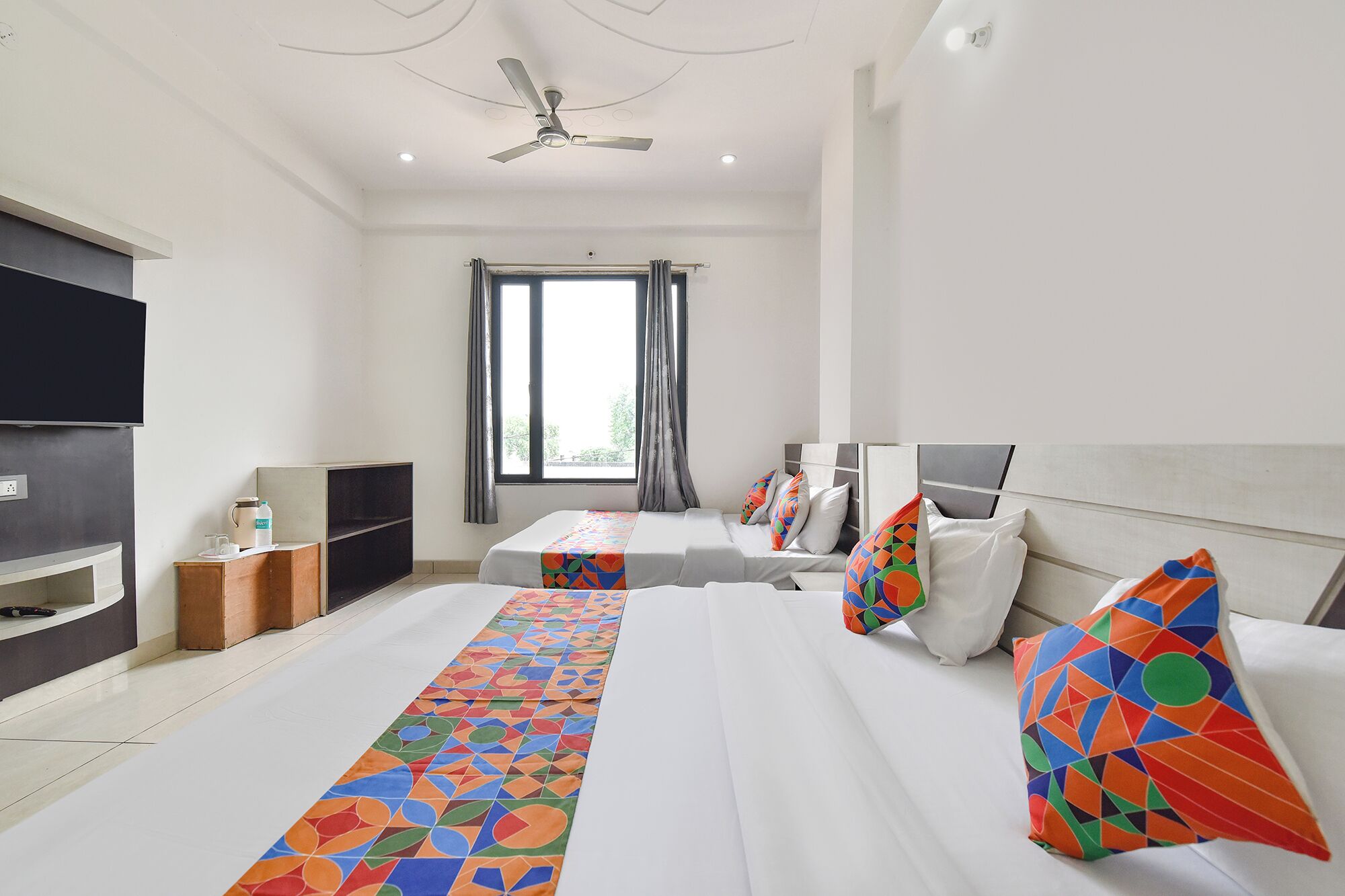 A hotel room with two beds, a TV, a ceiling fan, and a window with curtains.