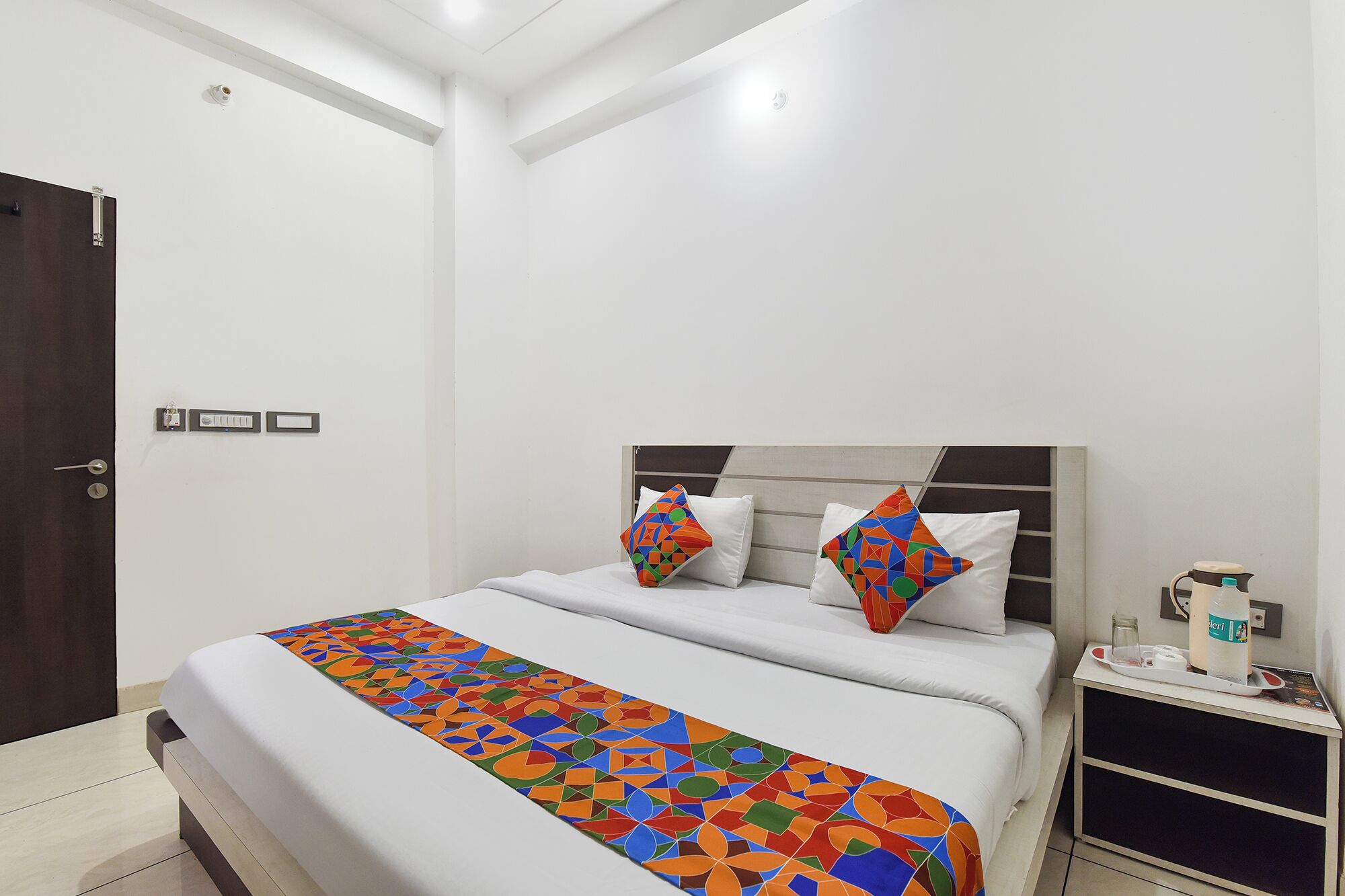 A hotel room with a bed, a bedside table with a lamp, and a door.