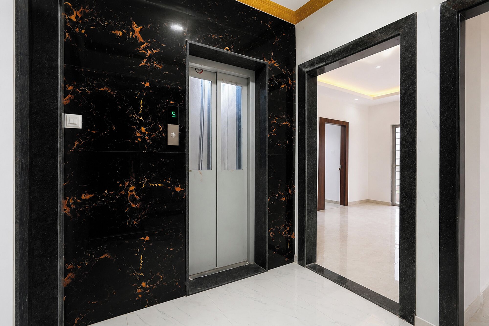 A hallway with a marble wall, a door with a digital display, and a mirror reflecting a room.