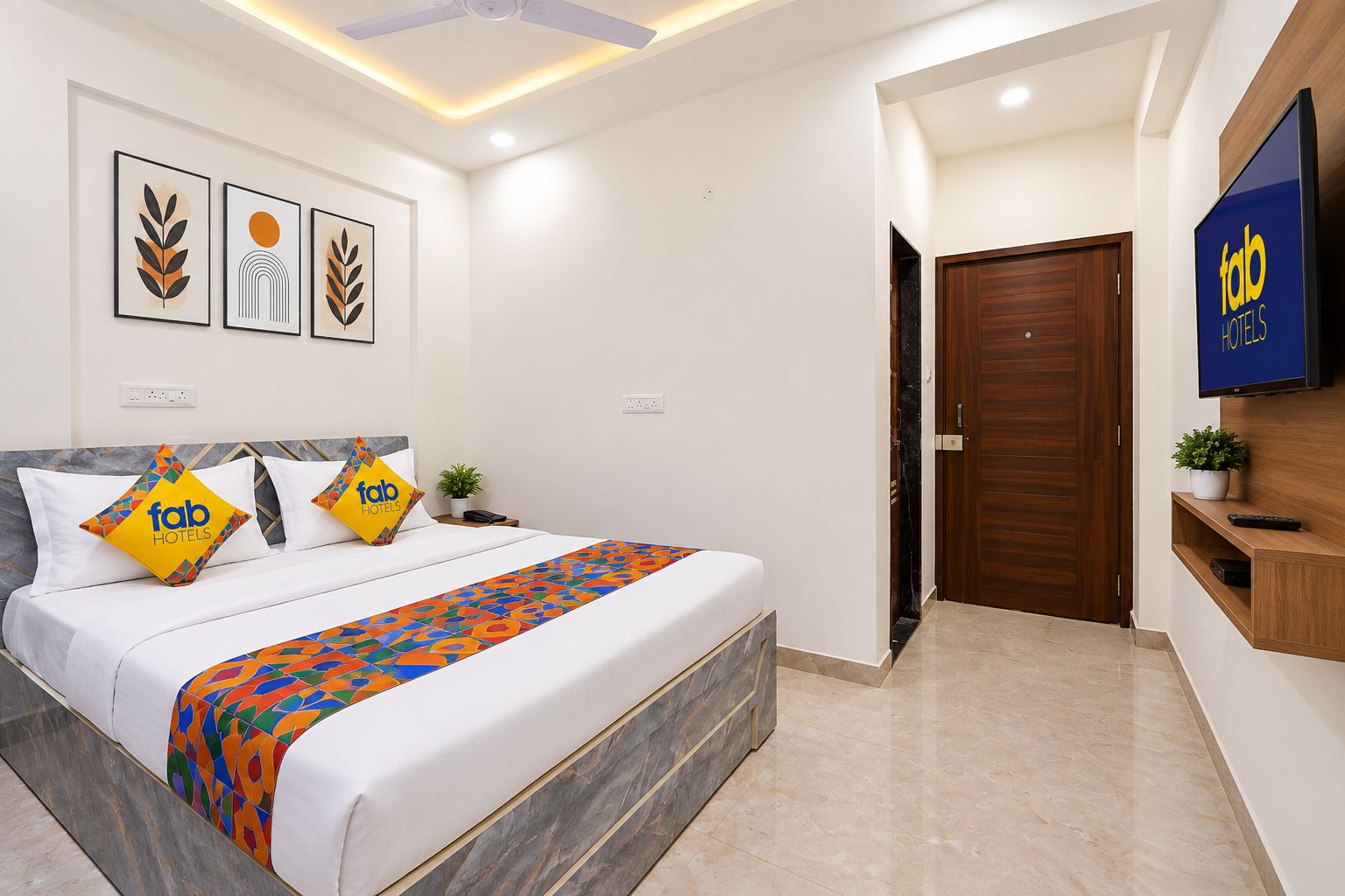 A hotel room with a bed, a TV, a door, and artwork on the wall.
