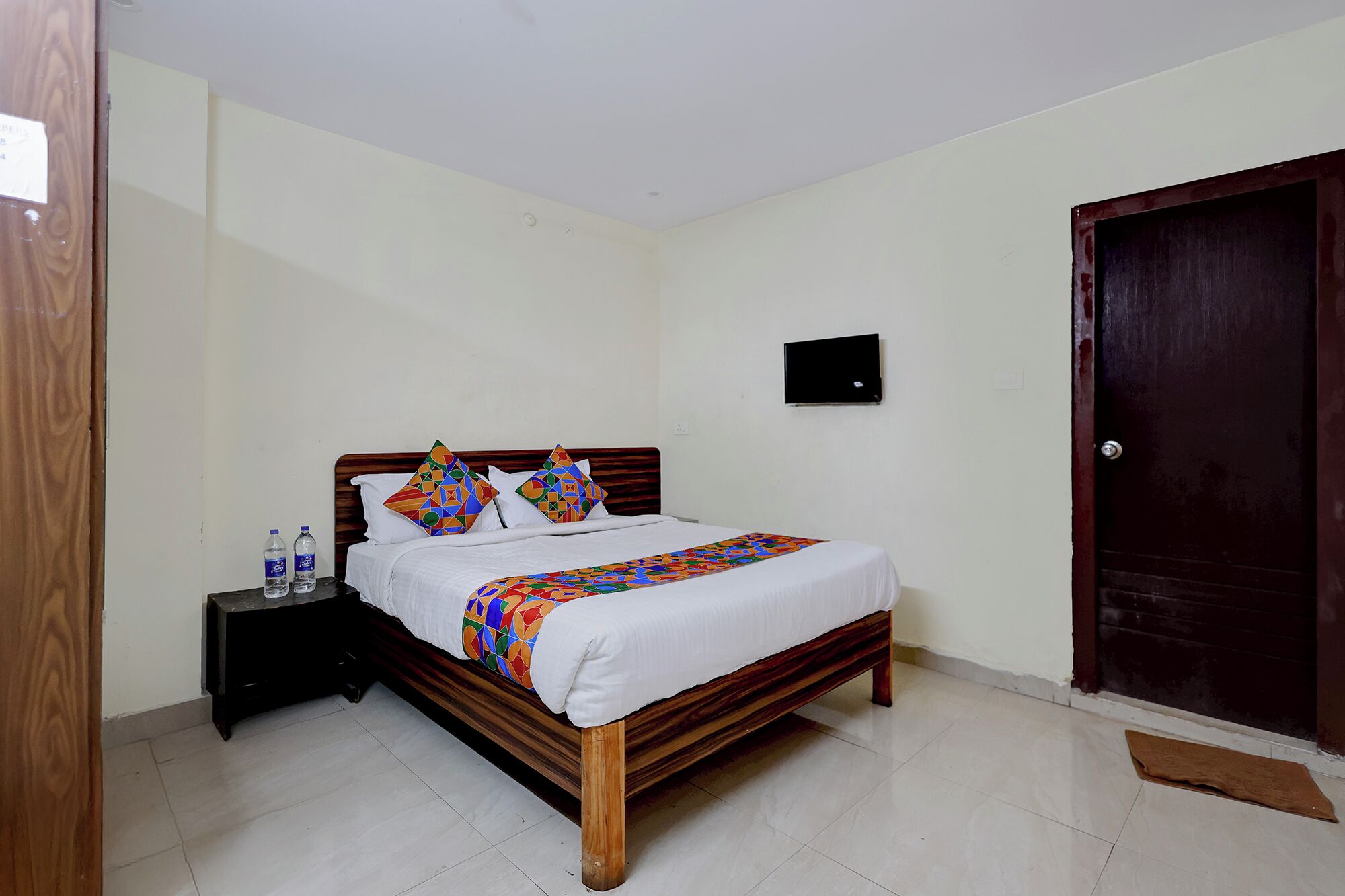 A bedroom with a wooden bed, white bedding, and colorful pillows. There is a small black TV on the wall and a wooden door.
