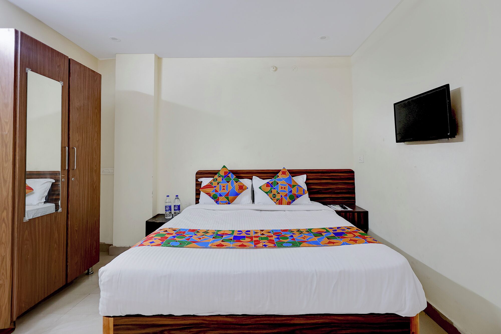 A hotel room with a bed, a wooden wardrobe, a nightstand, a television, and a mirror.