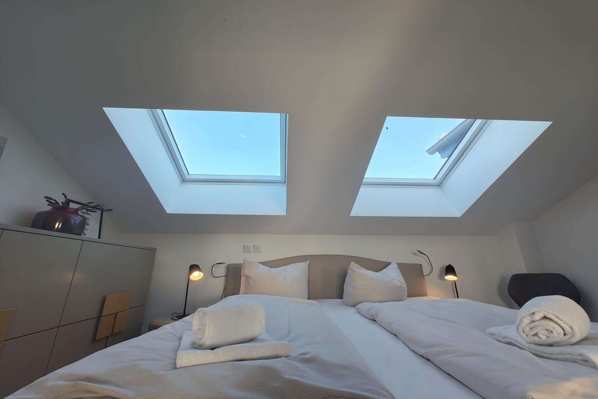 Ein Schlafzimmer mit einem Bett, zwei Oberlichtern, einem Nachttisch und einer Lampe.