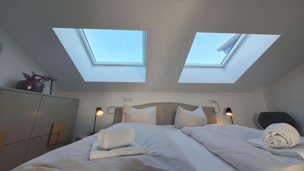 Een slaapkamer met een bed, twee dakramen, een nachtkastje en een lamp.