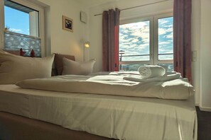 Ein ordentlich bezogenes Bett mit einem Kissen und einem gerollten Handtuch, ein Fenster mit Vorhängen und eine an der Wand befestigte Leuchte.