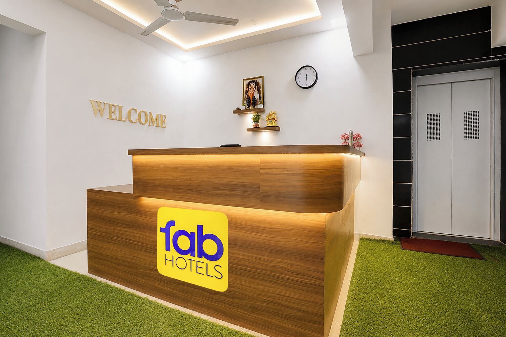 Eine Rezeption mit Holzoptik und dem Logo von 'fab hotels'. Über der Rezeption hängt ein Schild mit der Aufschrift 'WELCOME', und an der Wand befindet sich eine Uhr.