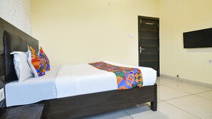A neatly made bed with a white sheet and colorful geometric-patterned blanket, a dark wooden headboard, and a plain wall with a door and a television mounted above it. - Fabhotel Shree I (Indore)