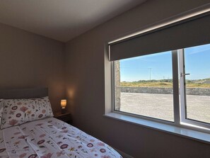 Ein Schlafzimmer mit Bett, Nachttisch, Lampe und Fenster mit Blick auf einen Parkplatz und die Ferne.