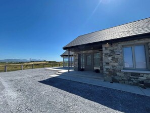 A modern stone building with a covered porch, large windows, and a gravel driveway. - Ballyconneely Lodge amazing views (Ballyconneely)
