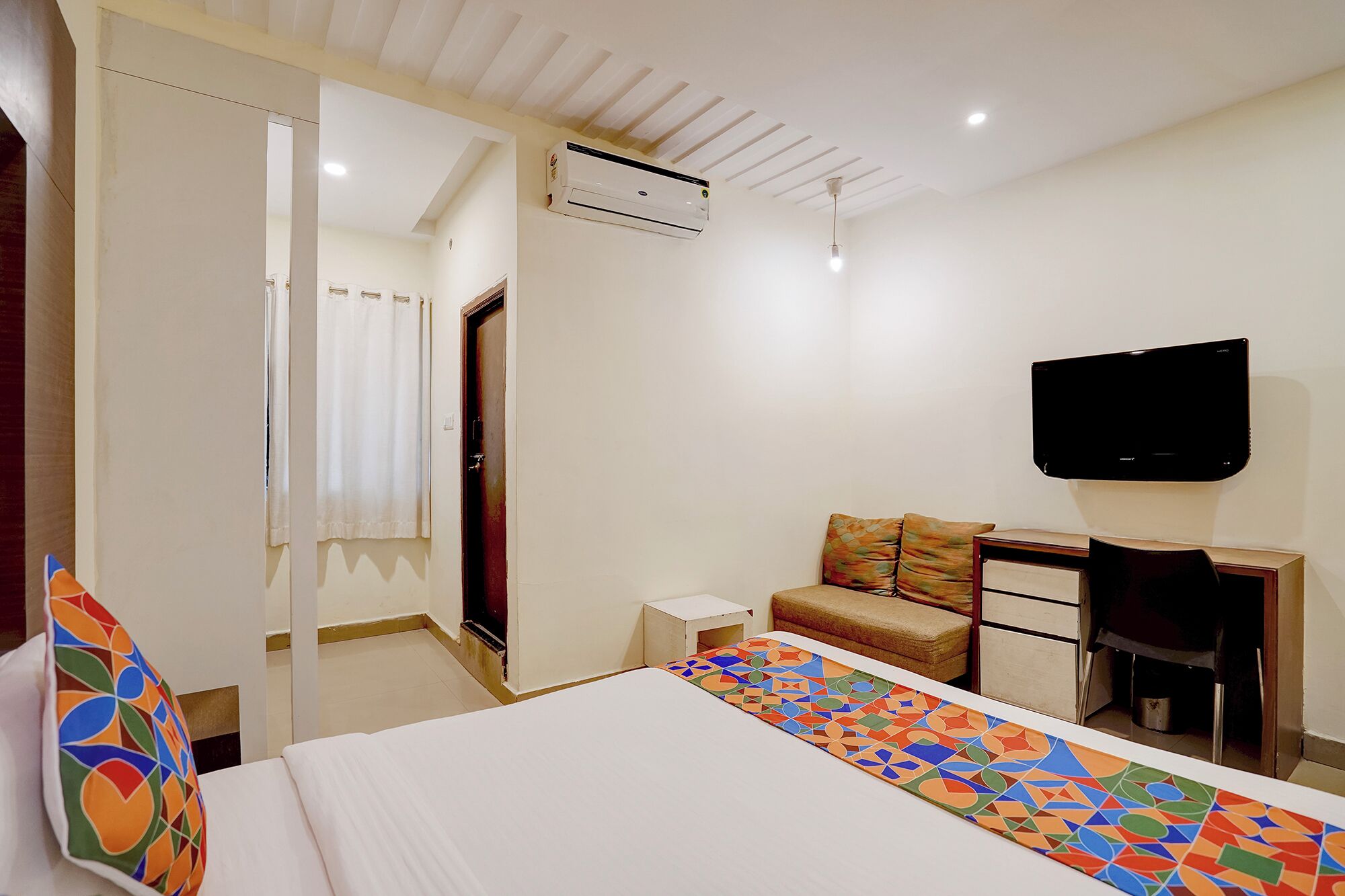 A hotel room with a bed, a television, a small table, a sofa, and an air conditioning unit.