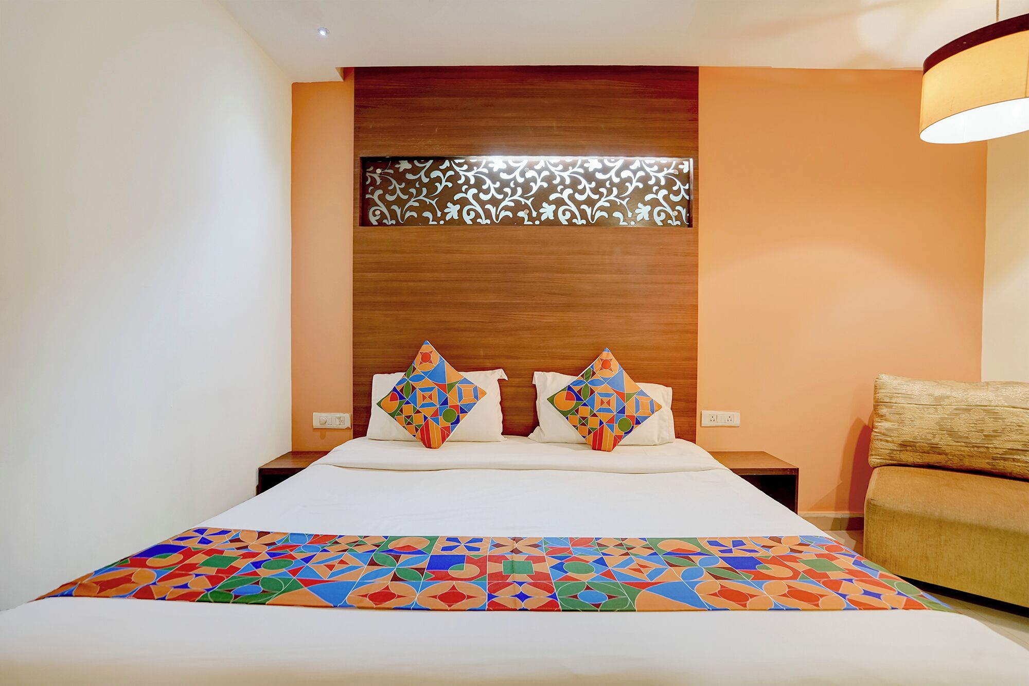 Quarto de hotel com cama, duas almofadas com padrões geométricos, mesa de cabeceira e luminária fixada na parede.